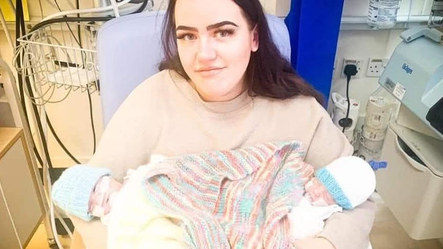 Meg Hughes, a woman with dark hair, smiles into the camera while holding new-born twins in her arms. The babies are wrapped in a rainbow-coloured blanket and each wearing woollen hats with tubes in their noses. The picture is taken on a hospital ward.