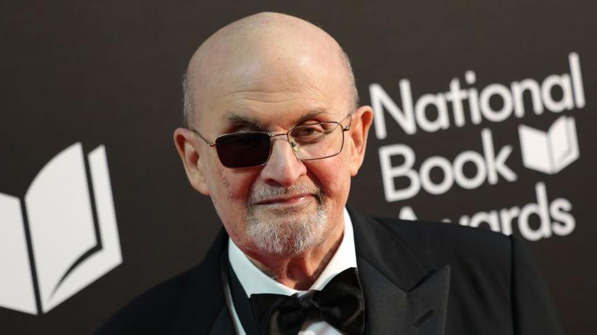 Salman Rushdi pictured at National Book Awards November 2024. He wears a black suit and black bow tie. He is bald and wears square glasses with one black lens and smiles at the camera.