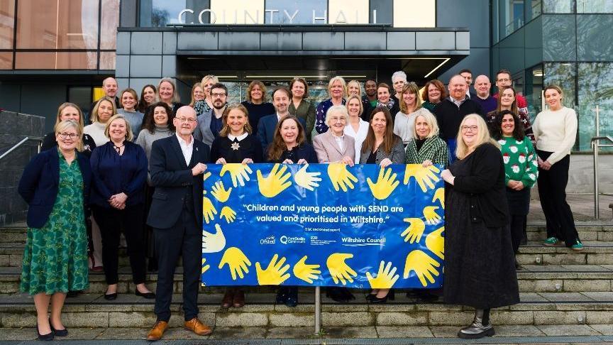 A group poses for a photo on steps outside the county hall. People in the front row are holding a poster reading Children and young people with SEND are valued and prioritised in Wiltshire. It is a blue poster with yellow hands around the sides.