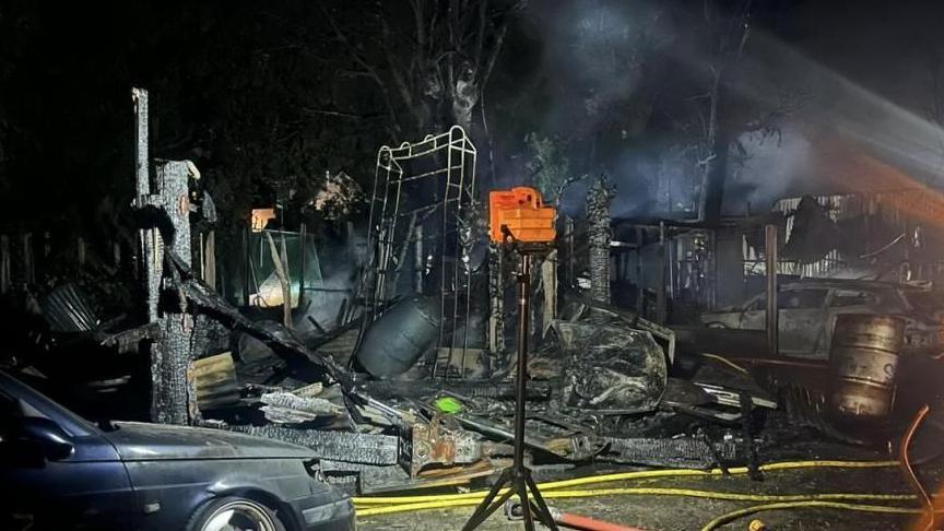 The blackened and charred remains of rubbish after the fire at night time. There are trees in the background.