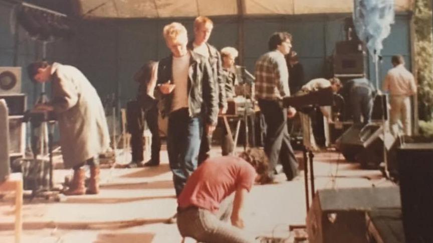 The band and crew are in various positions on stage in a tent during preparations for a gig.