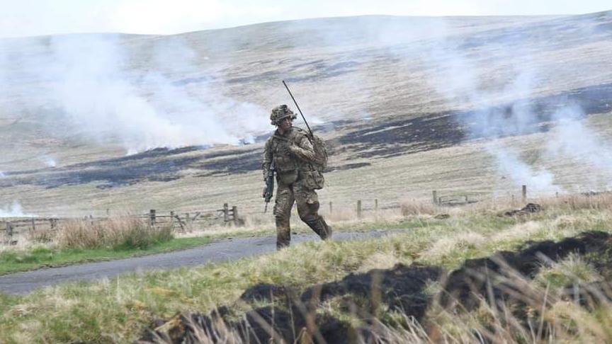 A soldier, wearing khakis, is walking through a hillside with smoke rising out of the grass.