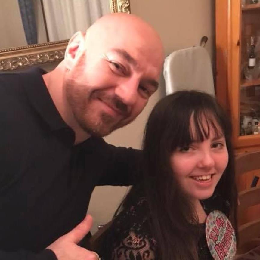 A bald man wearing a black long-sleeved top, gives a thumbs up with his right hand with his left arm around a girl, who has long dark hair, who is sitting down and smiling.