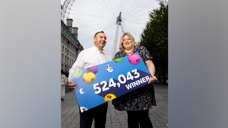Valdet Bujupi, wearing a white shirt, who has dark hair, holds a large blue cheque with the figure £524,043 on it and the wording 'winner.' On the right, is Abbey who is holding the other side of the cheque. They are standing in front of the London Eye.