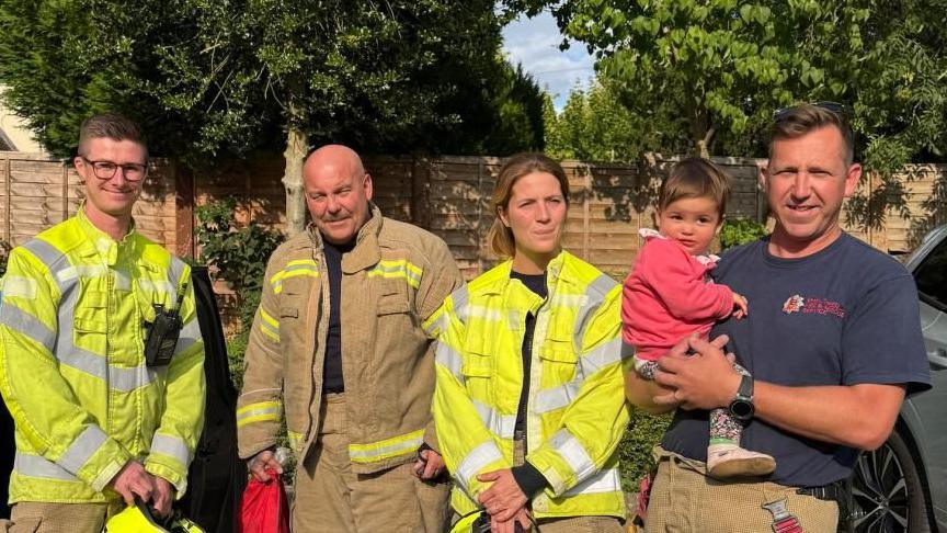 Four firefighters standing in a line, with the last one holding a girl in a pink top and floral leggings who looks to be around 18 months old