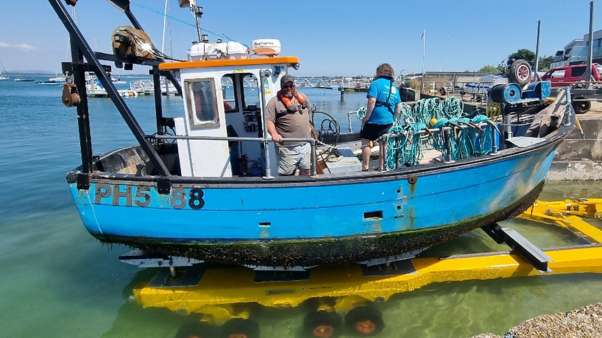 A small blue fishing boat, with two people on board, sits on a submerged yellow trolley on a slipway