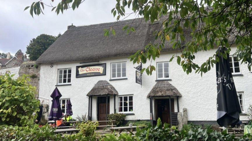 Outside The Cleave Inn in Lustleigh. The property is white with a thatched roof. Benches are outside the pub.