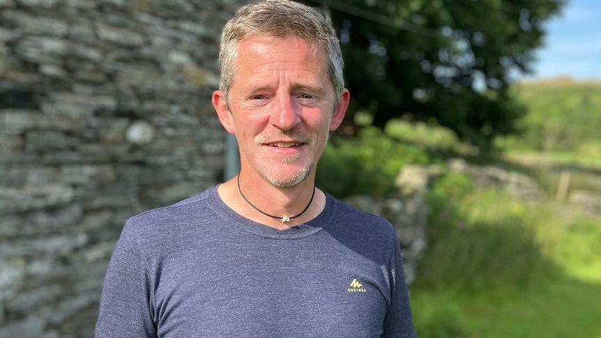 A man with short grey hair and some beard growth, wearing a blue t-shirt and standing outside a stone cottage