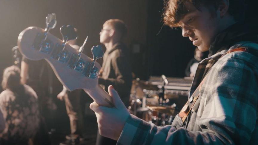 Man in a checked shirt playing a bass guitar in front of a drumkit on stage alongside other students singing into microphones