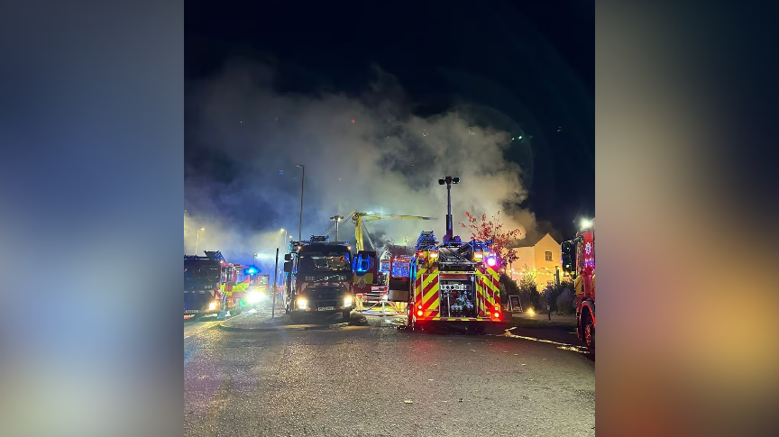 Fire engines can be seen parked on a street, pumping water into a building in the background that is on fire. It is night time and there is heavy smoke in the sky