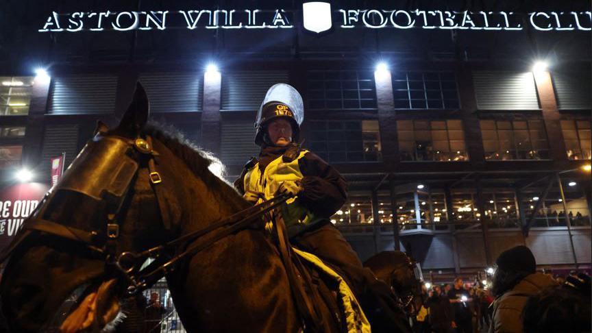 A police officer sits astride a horse which is in the foreground of the image, the ground is behind and the lit-up sign reading Aston Villa Football Club is in the background