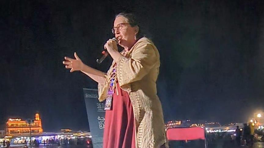 Lisa is shown holding a microphone and speaking during the event. It is dark, and the city lights are visible in the background. She is wearing a long red dress and a cream jacket, and has one hand up as she is talking. 