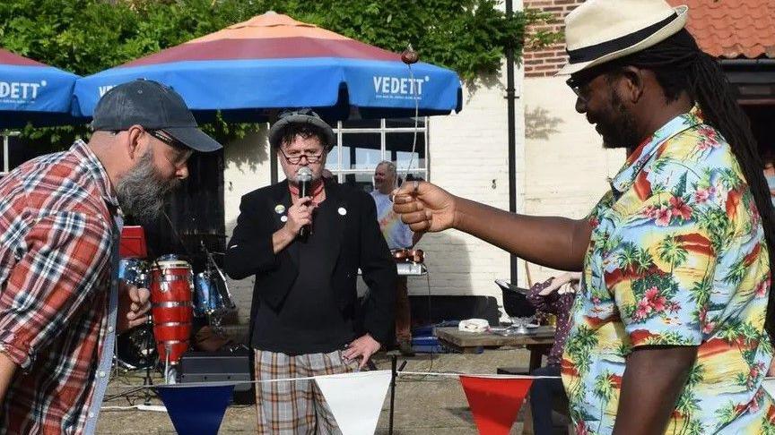 To men square up to play conkers as a third man in a hat stands in the middle with a microphone 