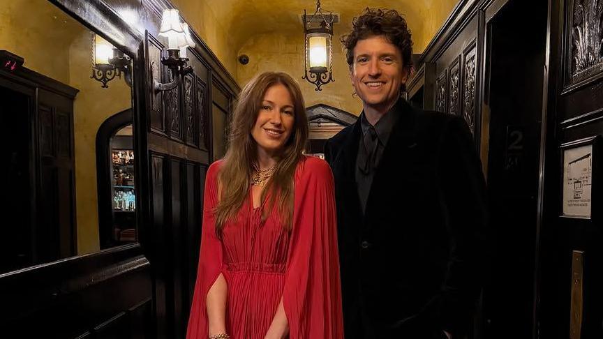 Bella wears a red long dress with cape and has long brown hair. Greg James wears a dark coloured suit with a grey tie and has short brown hair