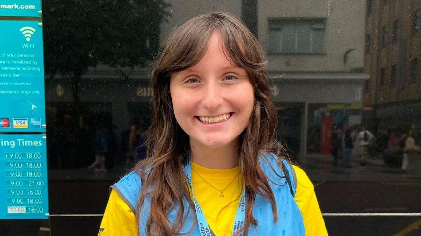A young woman, Addison McGowan pictured outside Primark home on Royal Avenue. She is wearing a yellow t-shirt with a blue bib on top and a blue lanyard. She has brunette hair.
