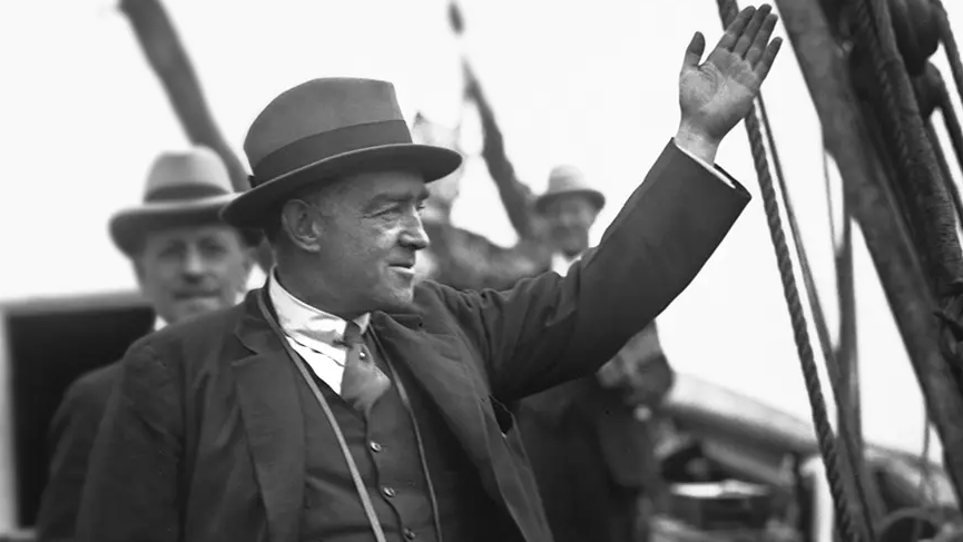 A black-and-white picture of Ernest Shackleton. He is standing on the deck of a ship wearing a hat, jacket, tie and a waistcoat. Two other men, wearing similar attire, can be seen in the background.