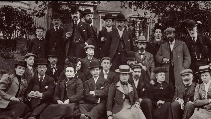 A group photo of the first members - male and female - of Halton Photographic Society wearing typical Victorian attire including straw boaters, top hats, bowler hats and flat caps.
