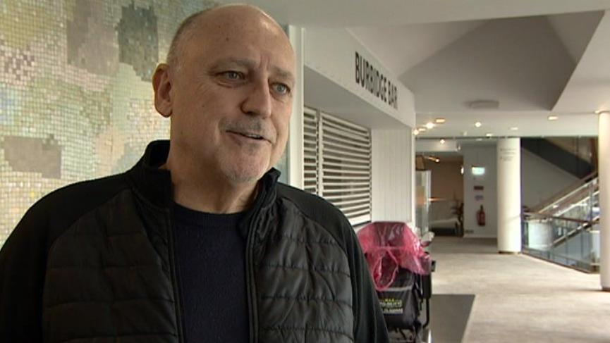 A bald man wearing a black jacket over a dark blue top standing in a corridor while speaking to someone off camera.