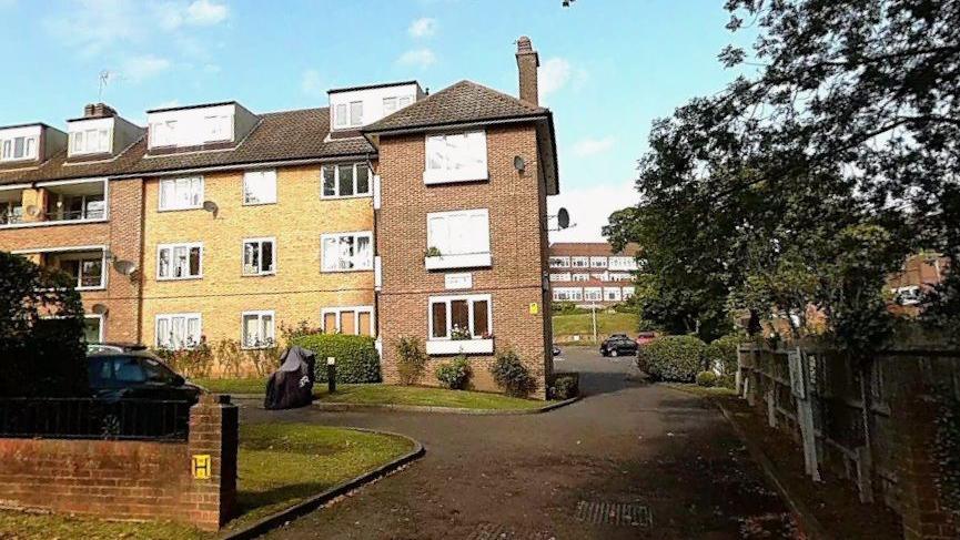 Google street map view of Kingfisher Court, off Queensmere Road, at three-storey apartment building with dormer windows and a paved driveway, bordered by grass, shrubs, parked vehicles and mature trees under a blue sky.