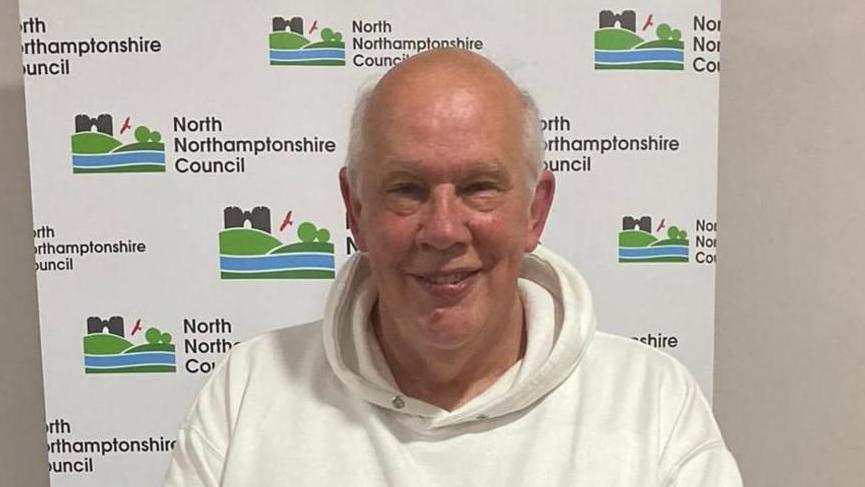James Hakewill with short white hair, smiling at the camera while wearing a white hoodie and sitting in front of a North Northamptonshire Council banner with its logo of trees in fields.