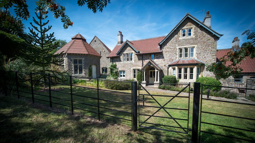 An exterior shot of the hospice which is a large grey and brown brick building, with red tiles on the roof. It is surrounded by a lawn and there are bushes and trees. In the foreground is a black metal fence and gate.