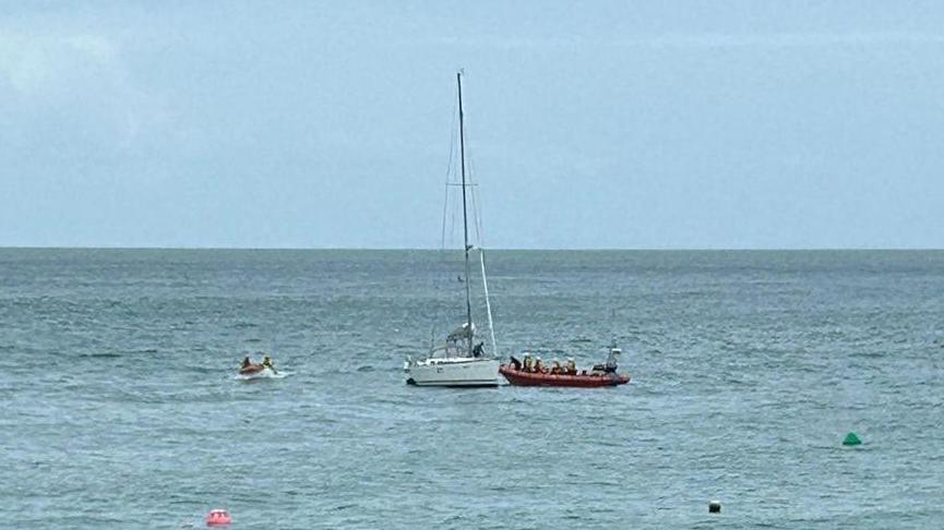 A small white yacht in blue seas. It is surrounded by two lifeboats. There are a couple of bouys dotted in the distance. The sky is blue. 