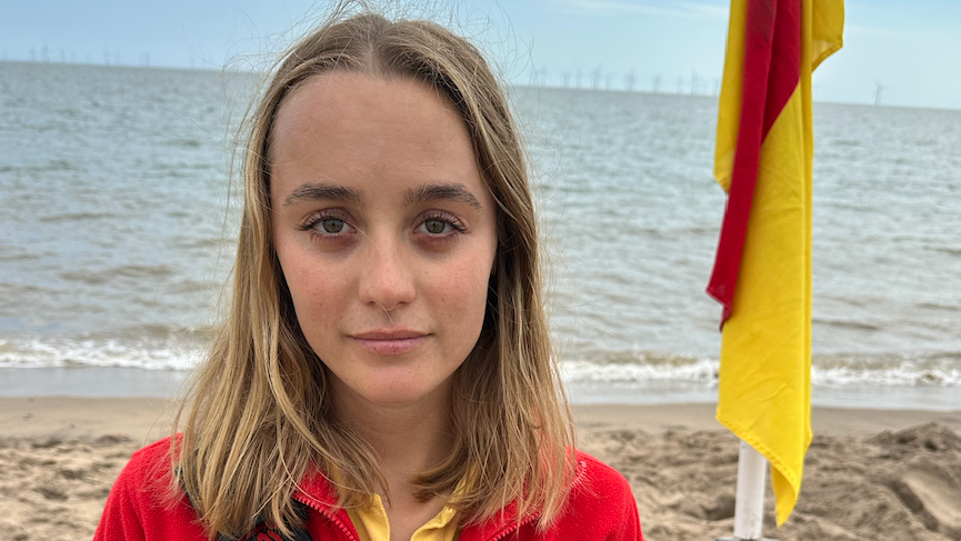 Lois Kemp is standing in front of the sea on the beach. She is wearing a red and yellow RNLI uniform. To the right of Lois is a red and yellow flag. Lois has blonde hair and blue eyes.