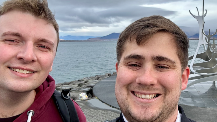 Elliot Griffiths and Zak Nelson facing the camera and smiling in a selfie in front an abstract metal sculpture with water and mountains in the background