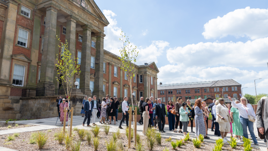 A side-on angle shot of dozens of smartly-dressed people at the ceremony outside a large brick building with stone pillars.