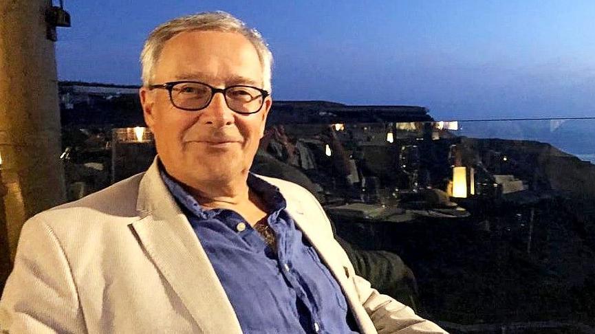 Head and body shot of Alex, 8th Marquess of Anglesey. He has short grey hair, wears dark-framed glasses and is smiling. He wears a blue shirt open at the collar with a light cream blazer open across the chest. He is sitting and the background is evening sky with reflected lights on a glass screened balcony.