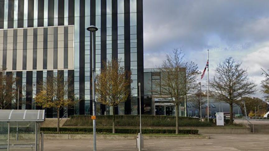 Tall, modern glass and steel building with single-storey entrance to the right. A flag is visible. There are trees around the building and a grey bus shelter to the left.