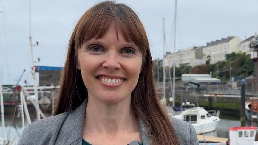 Joney Faragher, smiling: she has long straight brown hair with a fringe. She wears a green top under a grey jacket and stands in front of the quay in Douglas, with boats pictured behind her.