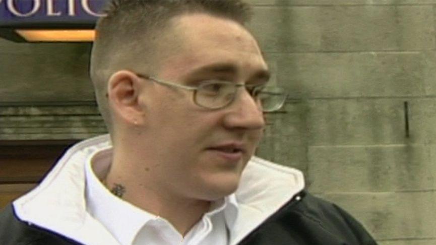 Old TV still of David Gibson wearing a white open-collar shirt and a black casual jacket. He has metal-rimmed glasses, crew-cut hair and a Chinese writing tattoo on his neck