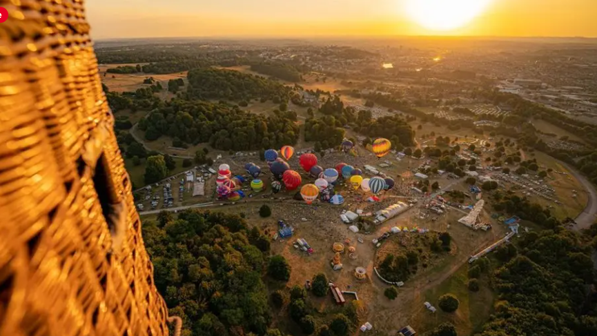 An aerial image taken from a hot air balloon basket, with part of it in view on the left. On the ground below is Ashton Court with lots of balloons being inflated and expansive views of the countryside at sunrise