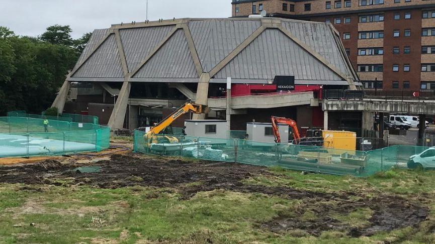 Hexagon theatre in Reading, with metal fencing around to secure the area for building. The image shows two diggers starting to dig up the earth around the theatre to install ground source heat pumps.