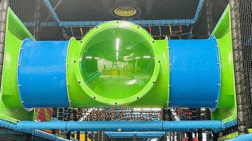 The image shows a section of a children's indoor playground. It shows a large cylindrical tunnel made of blue and green plastic, with a transparent green middle section. The tunnel is supported by blue padded bars and enclosed with safety netting. 