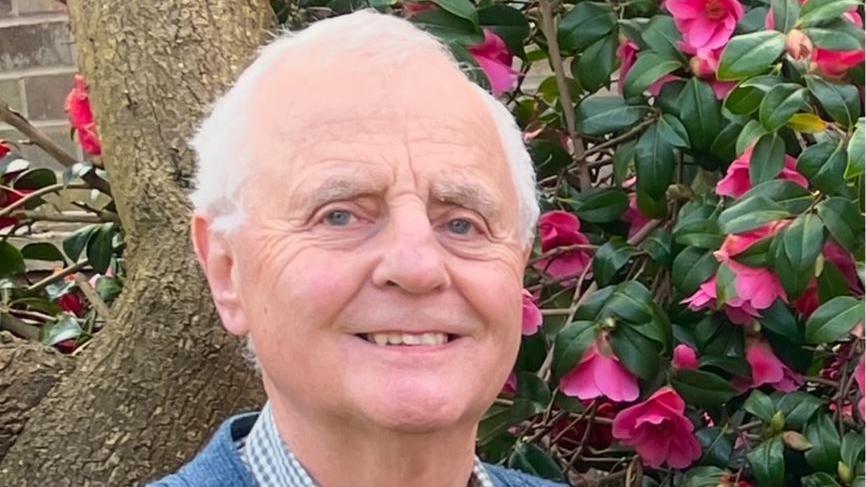 Peter Murphy wears a checked shirt and blue cardigan, standing next to a camellia plant with pink flowers