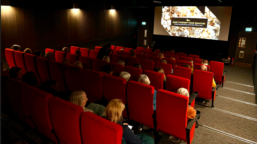 Rows of red seats make up a small cinema seating about 70 people. Most of the seats are taken and an advert is on the screen. 