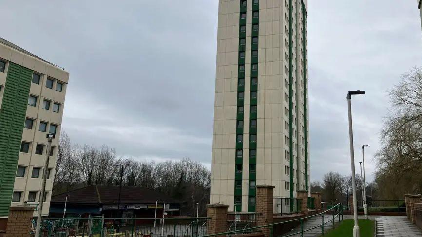 A tower block with green windows and beige cladding rises into the sky on a overcast day in Stockport