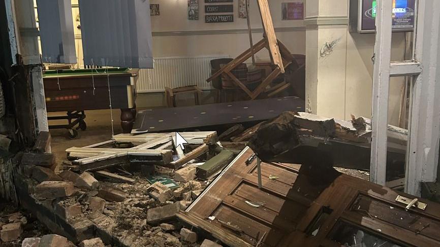 Broken bricks, rubble, and a broken wooden door lie in the middle of a bar area. A pool table can be seen undamaged