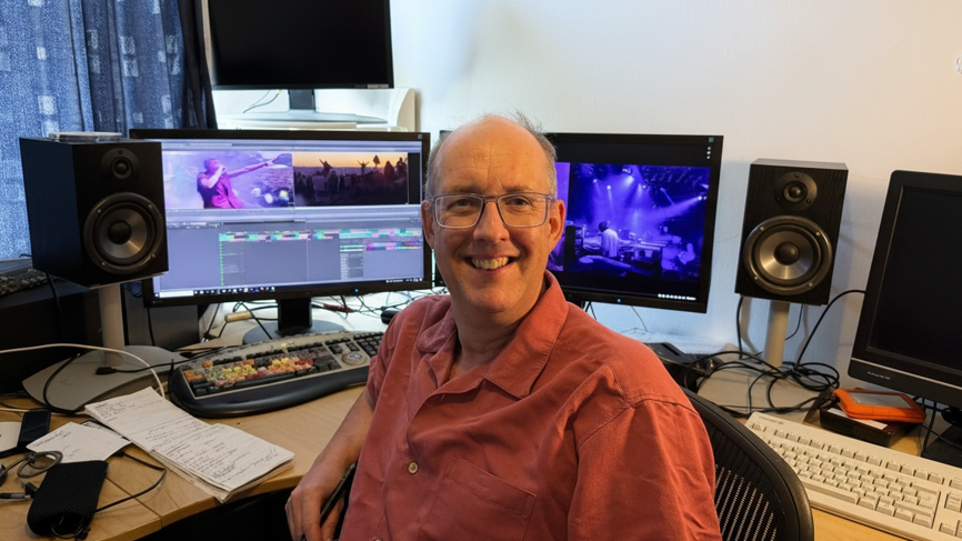 Robin sitting in a chair in an editing suite and smiling to camera. Behind him there are several monitors showing shots from the movie, and there are two large speakers either side. A curtain is drawn in the background
