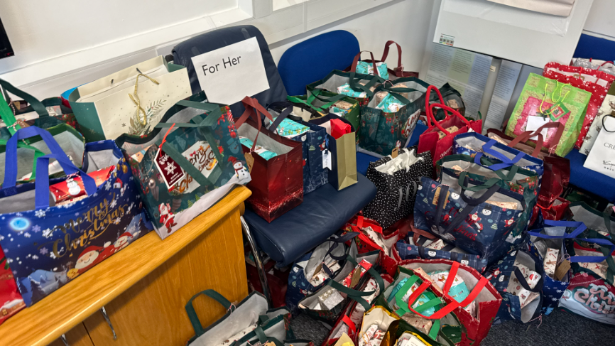 A number of gift bags containing presents are piled on a carpet. Bags have also been placed on chairs. The gifts bags are multicoloured. 