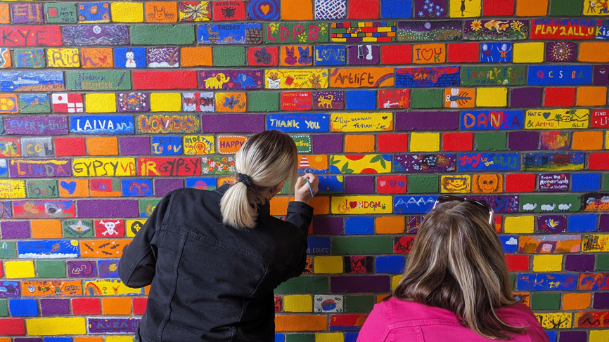 Two people with their backs to the camera each painting an image onto colourful bricks which form the mural in Gloucester.