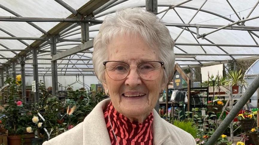 A lady wearing glasses and with short grey hair smiles . She is wearing a pink blouse and grey coat, and is standing in a garden centre, with flowers and plants behind her