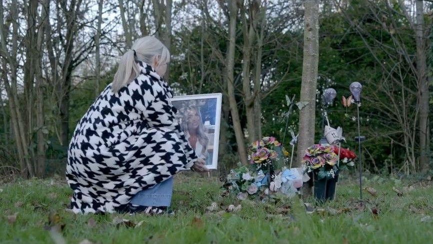 Ashley Dale's mother, Julie crouches down on grass near to trees to look at tributes to her daughter, including flowers. She is holding a large framed photo of her daughter.