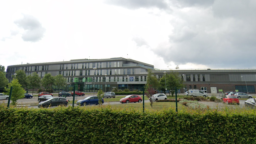 A Google streetview image of a school building which is set behind a green mesh fence and large green hedge. A number of cars are parked in front of the large grey building which features rectangular windows across three floors.