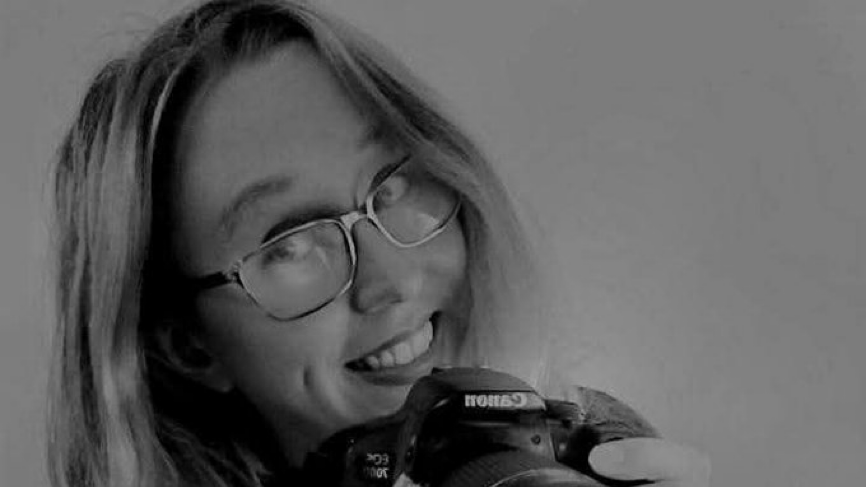 A black and white headshot of a lady, who is wearing glasses and smiling into the camera. She is holding a camera that reads "Canon". Her hair frames her face and is a light colour. 