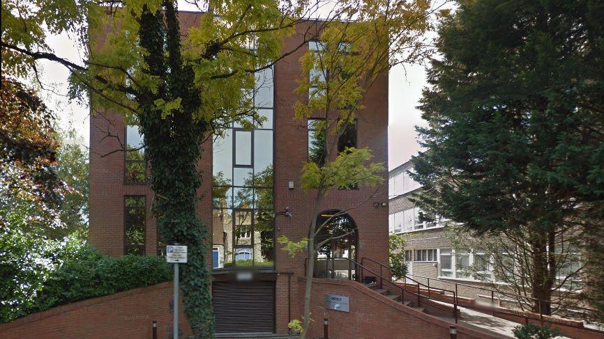 Three-storey brick office building with central glass section from top to bottom. A small parking area with a Senior sign is visible in the foreground. A grey office building is to the right.  There is a tree to the right and a hedge to the left.