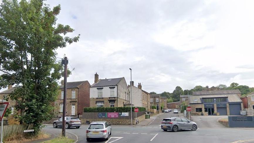 A shot of the junction between Church Lane and Bradford Road in Birstall - a T-junction with no entry signs flanking the road opposite and cars pulling out in both directions onto Bradford Road.