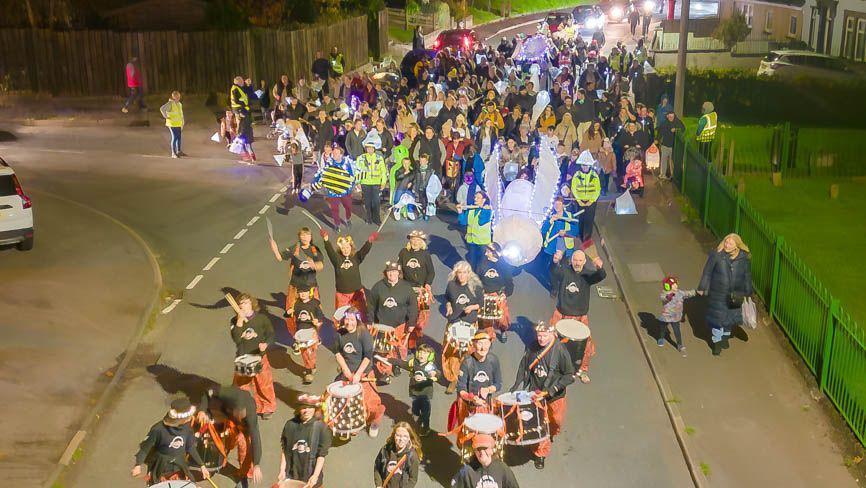 A large group of people fronted by a steel drum band walk thought the streets in Maryport holding lanterns and fairy lights